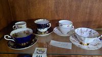 Five different teacups and matching saucers displayed on a shelf with varied floral and gold designs, colors include white, blue, and pink, note of a cracked saucer present.