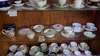 Wide shot showing multiple Royal Crown Derby teacups and saucers arranged on two glass shelves, some with blue tape indicating damage