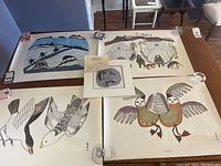 Four large Inuit prints on table including blue Arctic landscape, igloo scene, black & white birds, and colorful birds