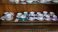 Photo showing a full view of 14 porcelain teacups and saucers with various floral patterns and some with gold trim displayed on a shelf.