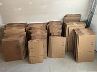 Photo showing stacks of flat cardboard moving boxes arranged by size, against a plain wall in a garage or similar space.