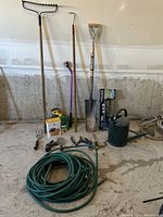 Photo of assorted garden tools including rake, shovel, watering can, hose, and small hand tools.