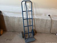 Blue P-handle hand truck dolly leaning against a concrete wall with visible floor and plumbing. The hand truck includes 4 flat cross bars and 2 black pneumatic wheels.