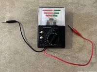 Front view of Radio Shack battery tester showing analog gauge, dial selector, and attached black and red test leads.