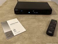Sony DVD/CD player with front panel display, remote control and printed manuals on table surface.