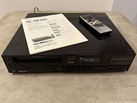 Front and top view of Sony SL-HF450 video cassette player and recorder showing digital display, buttons, and remote control resting on top alongside instruction manual.