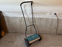 Photo of the Yardworks manual reel lawn mower showing the frame, comfort grip handles, teal reel, and black wheels on a concrete floor.