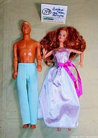 Full view of Barbie and Ken dolls together on light background. Barbie wears shiny gown with pink sash, Ken wears blue pants, no shirt.