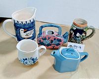 Photo showing five blue vintage ceramic pieces: a pitcher with a duck, floral creamer jug, pink/blue floral teapot, blue geometric teapot, and a ceramic stein with painted scene and lid.