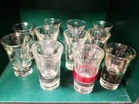 Ten assorted vintage clear glass shot glasses varying in size and shape, including one with a red band labeled 'Tsing Tao'