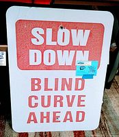 Photo showing front of vintage metal road sign with 'SLOW DOWN BLIND CURVE AHEAD' text and visible aging on red paint segments.