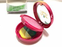 Opened compact showing rosy pink embossed blush and multicolored applicator sponge with box seen in background.