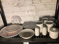 Overall view of various plates, cups, shakers, and sugar jar lined on a black metal shelf against a white brick wall.
