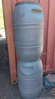 Two gray plastic 40 gallon rain barrels stacked vertically against a red metal wall.