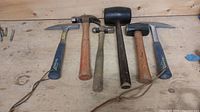 Full view of the six assorted hammers on a wood surface showing different types including pick, claw, ball peen, rubber mallet, and sledgehammer types.