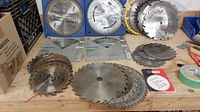 Full view of assorted saw blades spread on workbench showing different sizes and brands including packaged Hollow Ground Planet blades