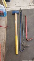 Photo showing sledge hammer with wooden handle and three breaking bars (red, blue, black) laid out on floor.