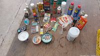 Overview of the lot with multiple automotive supply items arranged on the ground including lubricants, wax tins, spray cans, a grease tub, a tire gauge, and various small bottles.
