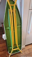 Full view of the underside of the vintage metal ironing board showing yellow legs, green metal surface with ventilation holes, and feet caps.