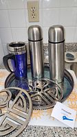 Two stainless steel Thermos brand coffee thermos bottles and one blue travel mug placed on a silver plated tray along with two zinc trivets.