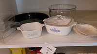Shelf with vintage Corningware dishes and plastic storage containers, including white ceramic dishes with blue floral and cornflower patterns, clear glass bowl, black tray, and stacked plates.