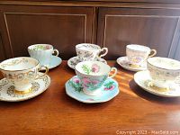 Six teacup and saucer sets arranged on wood surface showing different floral patterns and detailed craftsmanship.