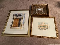 Three framed art pieces arranged on the floor; largest is a detailed watercolor door painting in white and gold frame, smaller ornate gold framed paintings depicting streets and buildings