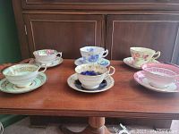 Six sets of china teacups and saucers arranged in two rows on wooden table, assorted floral designs and gold trim visible.