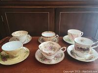 Photo showing six fine china teacup and saucer sets with various floral, scenic, and colored patterns, arranged on a wooden surface.