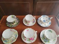 Photo showing six teacups on their matching saucers arranged on a wooden surface, displaying a variety of floral designs and patterns.
