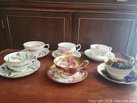 Six assorted fine china teacups paired with matching saucers, displaying various floral designs and gold accents.