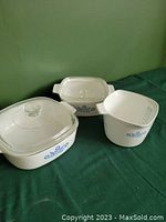 Three white Corningware pieces with blue cornflower design: one square saucepot and two lidded casseroles, side view.