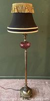 Full view of the floor lamp with black shade and gold fringe showing the brass stem and cranberry glass detail.