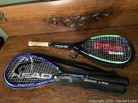 Wilson Defender and HEAD PP180 Graphite tennis rackets laid on wooden surface with Wilson in carrying case and strings visible, small red accessory (possibly tennis balls or overgrip) placed nearby.