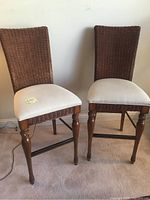 Front and side view of two bar stools showing woven rattan backs, fabric seats, and wooden legs with carved details.
