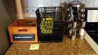Wooden box with 'Happy Birthday' label, two black wire baskets with handles, salt shaker and pepper mill on kitchen counter near stove.