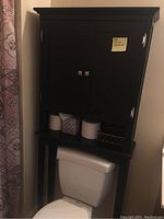 Full frontal photo of black over-the-toilet bathroom cabinet installed over toilet, showing doors, handles, and shelf area with bathroom items omitted in sale.