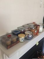 Twelve assorted scented candles in various containers, displayed on a white mantel.