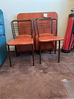 Photo showing four folding chairs with brown metal frames and rust-colored faux leather seats and backs, with a square card table featuring a wood-patterned laminate top behind them.