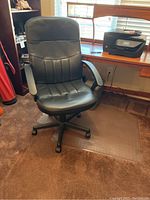 Black desk chair on brown carpet with armrests, caster wheel base, and vertical stitching on seat and backrest. Chair positioned near a wooden desk and printer.