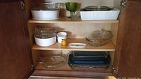 Interior cabinet shelf displaying various baking and serving trays including CorningWare French White rectangular, circular, oval dishes with lids and clear Pyrex casseroles.