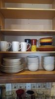 Cabinet shelves displaying white dinner plates stacked, white cereal bowls, colorful Fiesta plates and bowls in various colors (yellow, red, orange, blue) along with multiple mugs in white, dark blue, orange, and yellow.