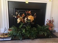 Five faux floral and plant arrangements placed on a fireplace hearth including leafy greens and autumn-colored flowers.