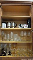 Glass cabinet with multiple shelves showing a variety of drinking glasses, metal travel mugs, plastic cups, and stemware.