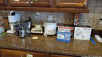 Photo showing several kitchen appliances including Krups coffee maker, Waring blender, Farberware crockpot, KitchenAid mixer boxes, Toastmaster chopper, Hamilton Beach grinder, NutriBullet shaker cups on kitchen counter.