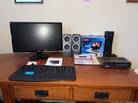 Lot displayed on wooden desk includes Acer monitor, keyboard, speakers, adapters, DVD player box, clock radio and desk light.