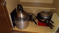 Photo of the left cabinet showing large stock pot with lid, stainless steel teapot on top, frying pan inside, and metal bacon tray at back corner.