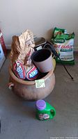 Photo showing 2 gardening pots, brown paper bag of Kingsford charcoal inside larger pot, handheld spreader, small fertilizer bottle in foreground.