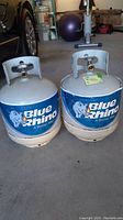 Two Blue Rhino propane tanks shown side by side on a garage floor with protective neck guards and Blue Rhino branding.
