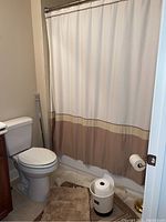 Shower curtain on rod above bathtub, bath mats visible on floor, white bathroom trash can beside toilet, toilet paper holder on wall-mounted dispenser